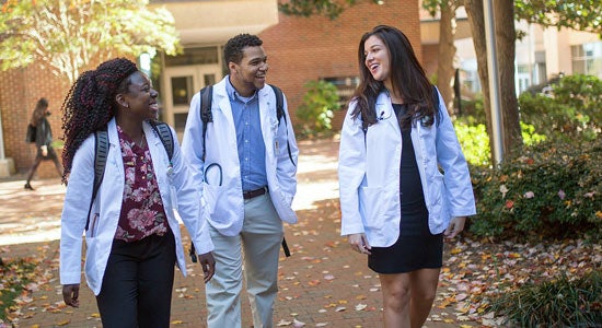 three medical students in white coats walking on campus together.