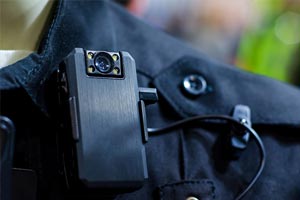 Close-up of a black body camera attached to a dark uniform, featuring a lens and visible wiring.