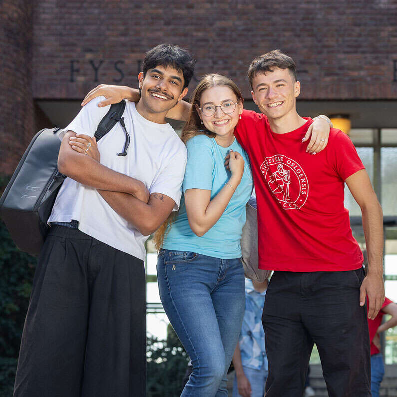 A group of three smiling students standing outside the Physics building with main entrance in the background.