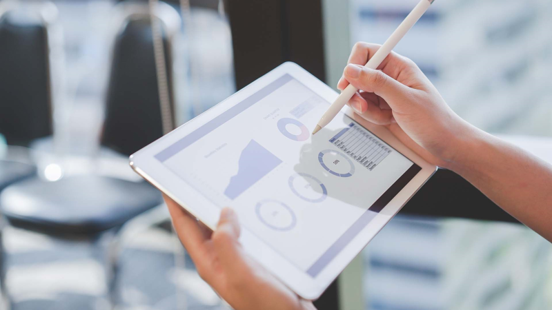 Close-up of a person's hands holding a tablet and using a stylus to interact with a digital analytics dashboard displayed on the screen. The dashboard shows various charts and graphs related to business performance. The background is softly blurred, showing a bright office environment with empty chairs, suggesting a setting of professional analysis or review.