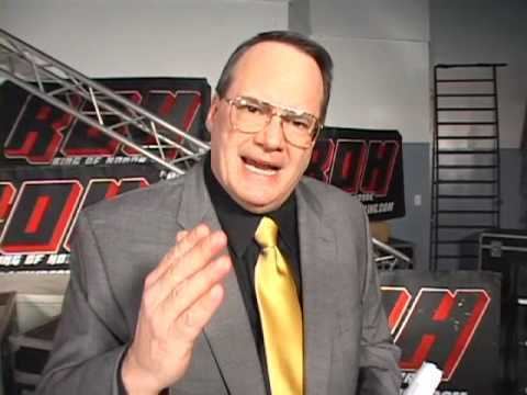 Jim Cornette with an angry expression, wearing eyeglasses, a gray suit, black long sleeves, and a golden yellow tie.