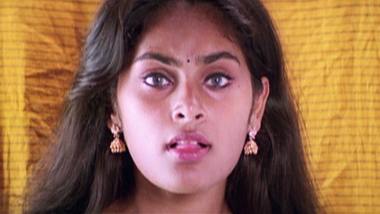 Shrutika with black hair and a serious face while her tongue is out and she is wearing gold earrings