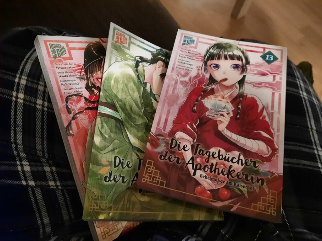 Drei Bände von "Die Tagebücher der Apothekerin" (genau genommen Band 13, 14 und 15), die aufgefächert auf meinen Beinen liegen.