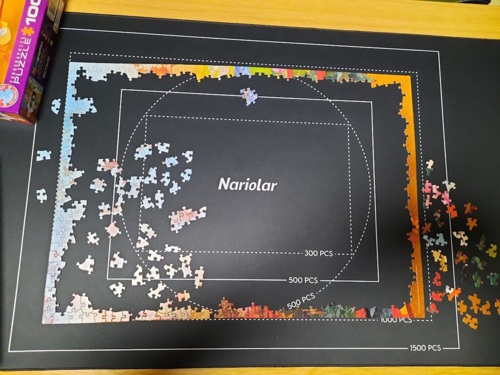 Ein angefangenes Puzzle auf einer Matte. Der Rahmen ist fast vollständig und auf der linken Seiten im Rahmen liegen einige hellblaue und rosa Puzzleteile.