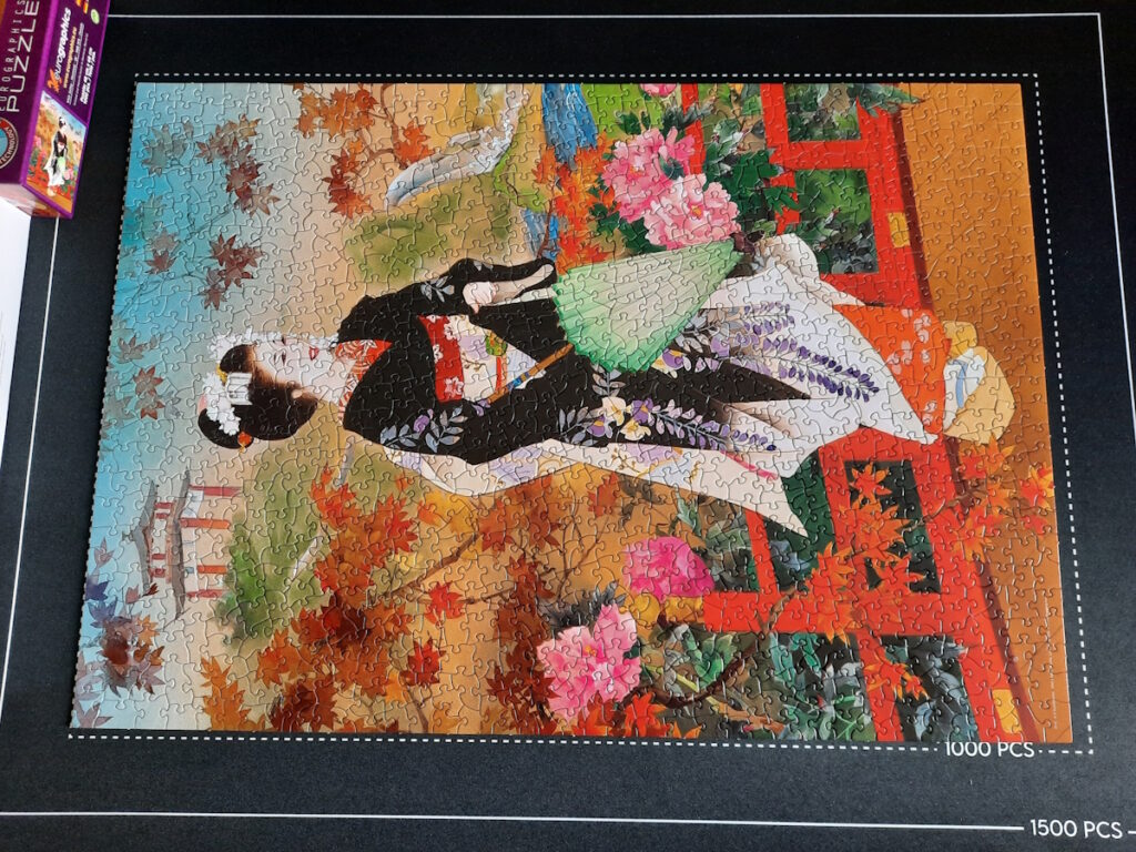 Ein fertigs Puzzle, dessen Motiv eine Geisha in einem schwarz-weißem Kimono mit Blütenmuster zeigt. Die Geisha steht mit einem hellgrünen Schirm in der Hand auf einer roten Brücke. Im Hintergrund ist ein Tempel zu erahnen, direkt hinterm Brückengeländer sind blühende Bäume zu sehen.