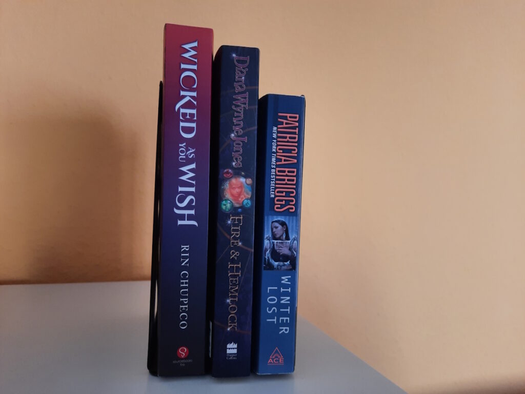 Drei Bücher vor einer orangen Wand. Die Titel der Bücher lauten von links nach rechts: Wicked as You Wish von Rin Chupeco, Fire and Hemlock von Diana Wynne Jones und Winter Lost von Patricia Briggs.