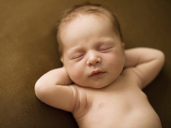 Sleeping newborn baby lying on a brown surface with arms behind head.