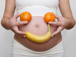 Pregnant belly with hands holding oranges and banana forming a smiley face.