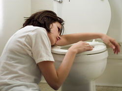 A young person in a white shirt leans over a toilet, appearing sick or nauseous in a bathroom.