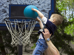A young child is being lifted by an adult to shoot a blue basketball into an outdoor hoop.