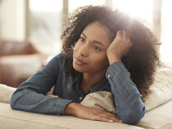 A woman with curly hair rests her head on her hand, looking thoughtfully into the distance while leaning on a beige couch in a softly lit living room.