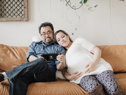 A couple relaxes on a tan couch, the woman pregnant, playing a Nintendo Switch together.