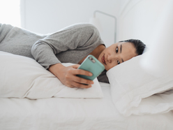 A woman lies in bed looking at her smartphone, appearing thoughtful and relaxed.