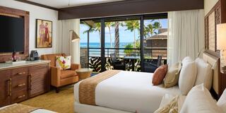 A stylish hotel room features two beds, a cozy chair, a flat-screen TV, and a stunning balcony view of palm trees and ocean waves.