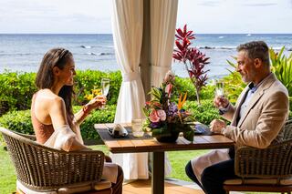 A couple enjoys a toast with champagne at a beautifully set table by the ocean, surrounded by lush greenery and vibrant flowers.