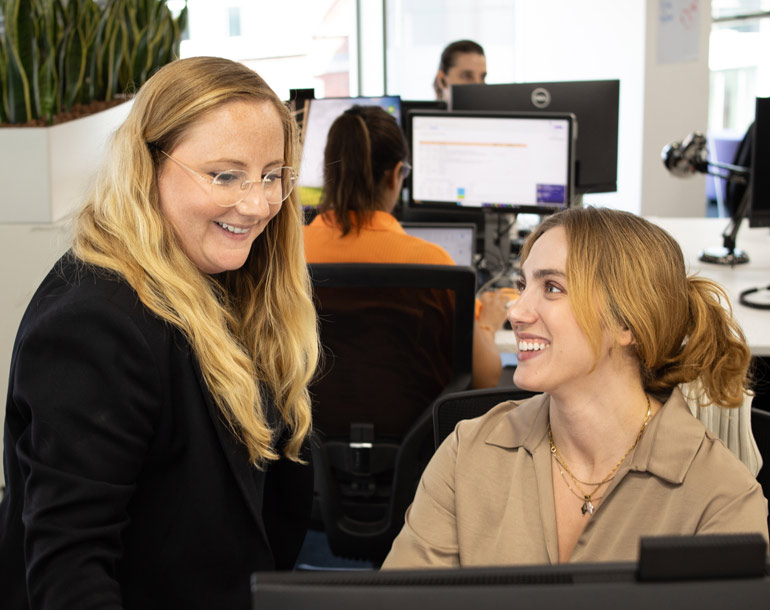 Two women smile and talk at a computer desk in an office setting. Another person works at a computer in the background.