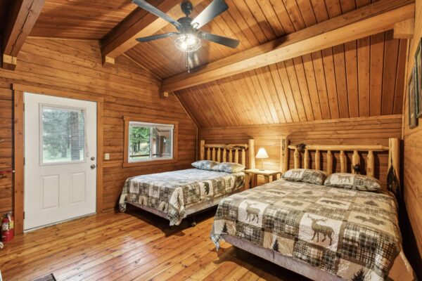 Wood-paneled cabin bedroom with two double beds, rustic bedding, a bedside lamp, ceiling fan, wooden floor, and a window and door looking out to trees.