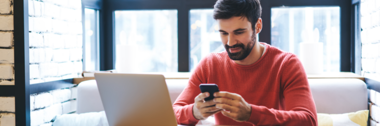 Man in red sweater using smartphone with laptop on table in a modern room with large windows.