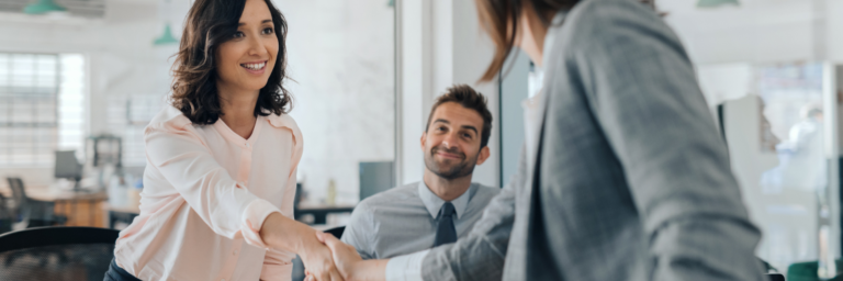 Two professionals shaking hands during a meeting with a colleague observing.