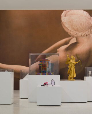 Gallery with large artwork of a reclining figure on the wall. Foreground features modern sculptures on white pedestals, including a golden figure. Traditional paintings frame the scene.