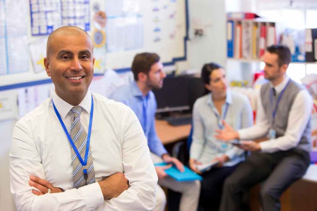 Smiling school administrator standing with arms crossed while a group of educators meet and discuss in the background.