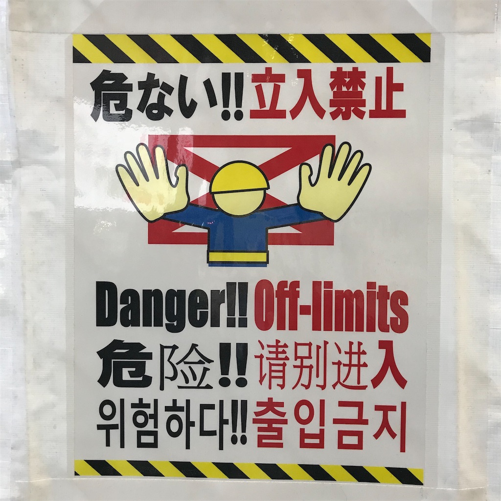 両腕を広げた顔のないヘルメットの人のイラスト、上下に黒と黄色の斜め縞のついた貼り紙、白いガムテープで貼られている。日本・英語・簡体中国語・ハングルの4言語表記。