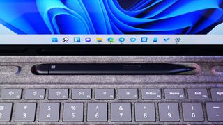 A Surface Slim Pen 2 rests in a recessed slot above a grey keyboard, with a tablet screen displaying the Windows 11 taskbar in the background.
