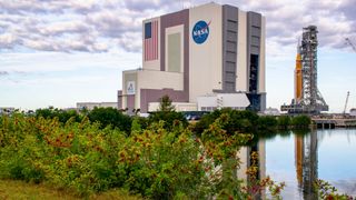 An orange rocket sits on a mobile scaffold as it rolls out of a large white building with the American flag and NASA meatball logo on the side.