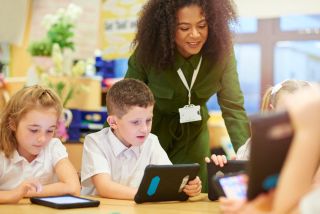 Four elementary school kids working on tablet computers while smiling teacher watches.