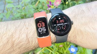 The Fitbit Charge 6 on an orange strap next to the Pixel Watch 3 on a black strap on a user's wrist with blue and pink flowers in the background