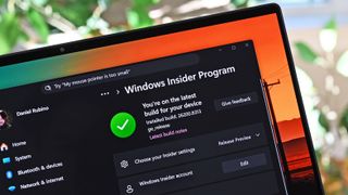 Close-up of a laptop screen displaying the Windows Insider Program page. A green checkmark indicates the latest build is installed. The background shows blurred greenery outside.