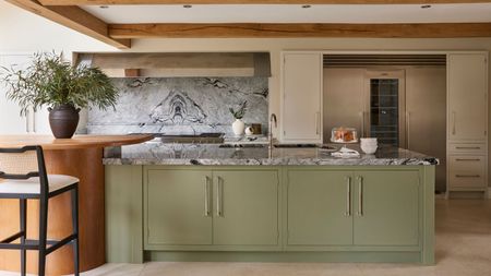 A kitchen with a large green painted kitchen island and marble countertop, built into a large orange kitchen table with black bar stools. A floor-to-ceiling fridge freezer unit built in in the background.