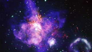 a highly detailed composite image of a star-forming region of space known as 30 Doradus, shaped like a bouquet, or a maple leaf. 30 Doradus is a powerful stellar nursery. In 23 days of observation, the Chandra X-ray telescope revealed thousands of distinct star systems. Chandra data also revealed a diffuse X-ray glow from winds blowing off giant stars, and X-ray gas expelled by exploding stars, or supernovas. In this image, the X-ray wind and gas takes the shape of a massive purple and pink bouquet with an extended central flower, or perhaps a leaf from a maple tree. The hazy, mottled shape occupies much of the image, positioned just to our left of center, tilted slightly to our left. Inside the purple and pink gas and wind cloud are red and orange veins, and pockets of bright white light. The pockets of white light represent clusters of young stars. One cluster at the heart of 30 Doradus houses the most massive stars astronomers have ever found. The hazy purple and pink bouquet is surrounded by glowing dots of green, white, orange, and red. A second mottled purple cloud shape, which resembles a ring of smoke, sits in our lower righthand corner.