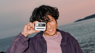 Young man happily holding a Polaroid Go Gen 3 tiny instant camera to his eye, with the sea and a sunset visible in the background