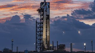 A black and white SpaceX Falcon 9 rocket carrying the Ax-4 crew capsule on the launch pad with colorful clouds lit by twilight.