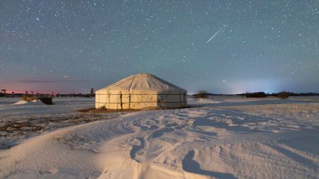 A meteor streaks throught the starry night sky above a fabric structure surrounded by snow.