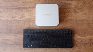 A white Geekom A9 Max mini-PC placed on a wooden desk above a black Razer Joro keyboard.