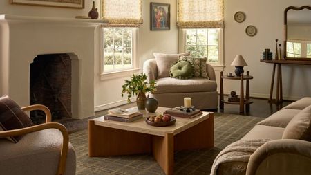 A cozy but bright cream living room with cream upholstered chairs, cuddle seats and mid-centrury wooden coffee table, side table and sideboard. There is a stone mantel and sheer window treatments in view, as well as a checkered area rug