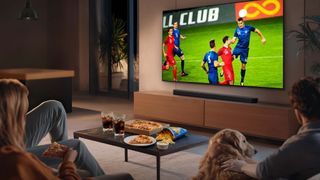 A lifestyle image showing a couple with a dog watching football on a Samsung QN80H. The image was in part created with AI
