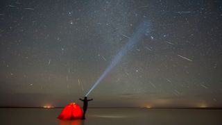 A person stands with arms outstretched behind a bright orange tent looking up at the night sky full of streaks with meteors with a large flashlight beam pointed up.