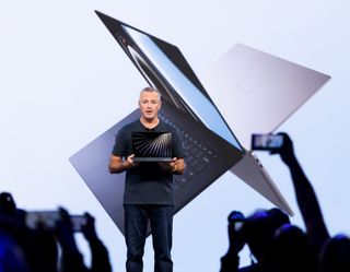 Man presenting a sleek laptop on stage, with a large image of the device projected behind him. Audience members capture the moment on phones.