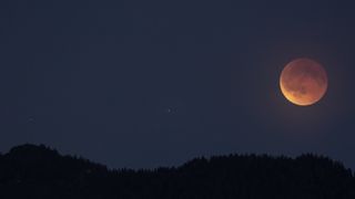 A orange glowing moon is seen against a dark blue night sky in the right side of the image with a silhouetted forested hillside below.