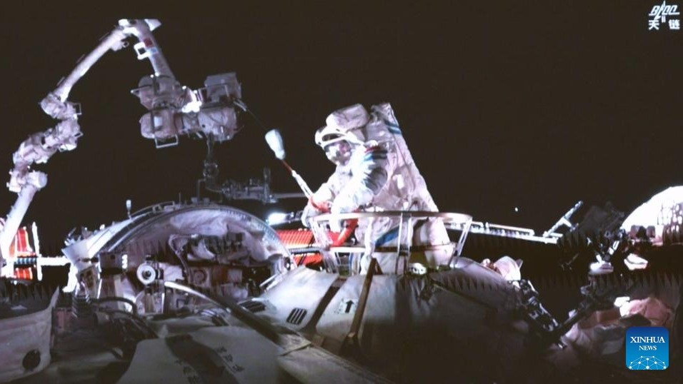 an astronaut rises from the hatch of a circular space station node. A robotic arms stands in the distance on the elft.