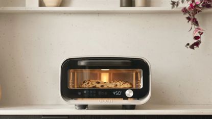 Ooni's volt 2 indoor pizza in poplar white cooking a pizza inside on top of a kitchen counter underneath open shelving with decor items