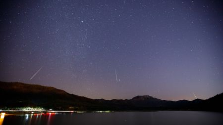 White streaks of meteors are seen against a dark purple night sky with a frozen lake with lights below pushed against silhouetted mountains in the background