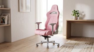 Sleek pink gaming chair with armrests in a sunlit room, set on a wooden floor with a plant on a wooden desk nearby, conveying comfort and style.