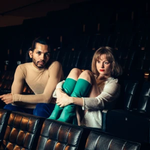 Man and woman sitting apart in a theater with empty black seats around them.