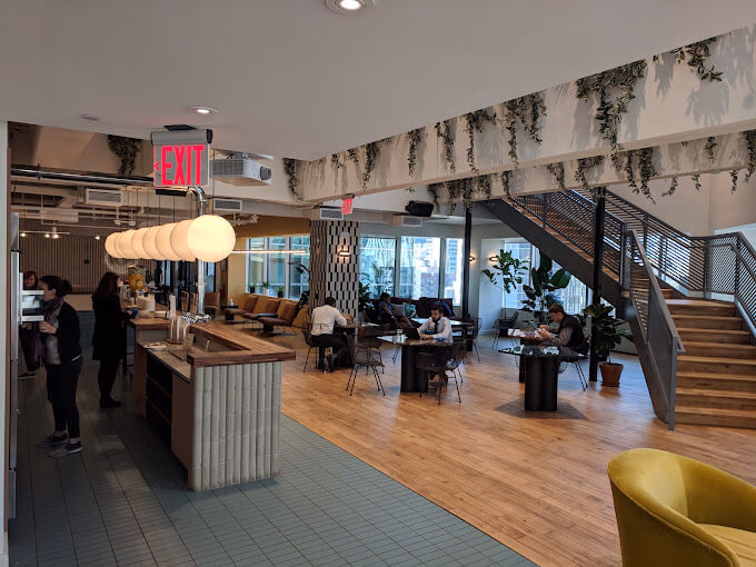 Modern coworking space with people sitting at tables. Features wooden floors, greenery, and decorative lights. A welcoming, collaborative atmosphere.