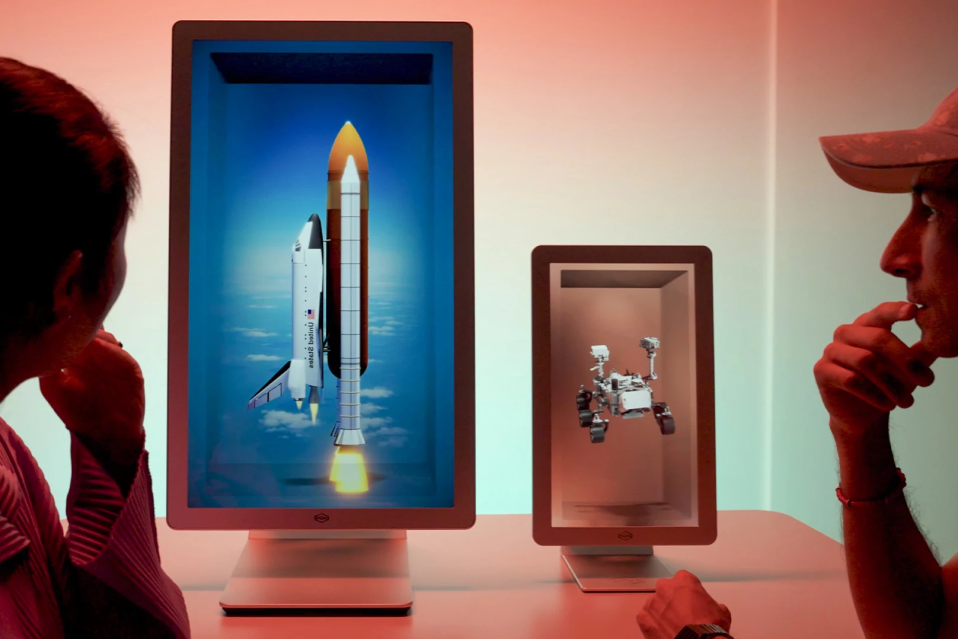 Two people observing two Hololuminescent™ Display (HLD) showing a space shuttle launching and a Mars rover.