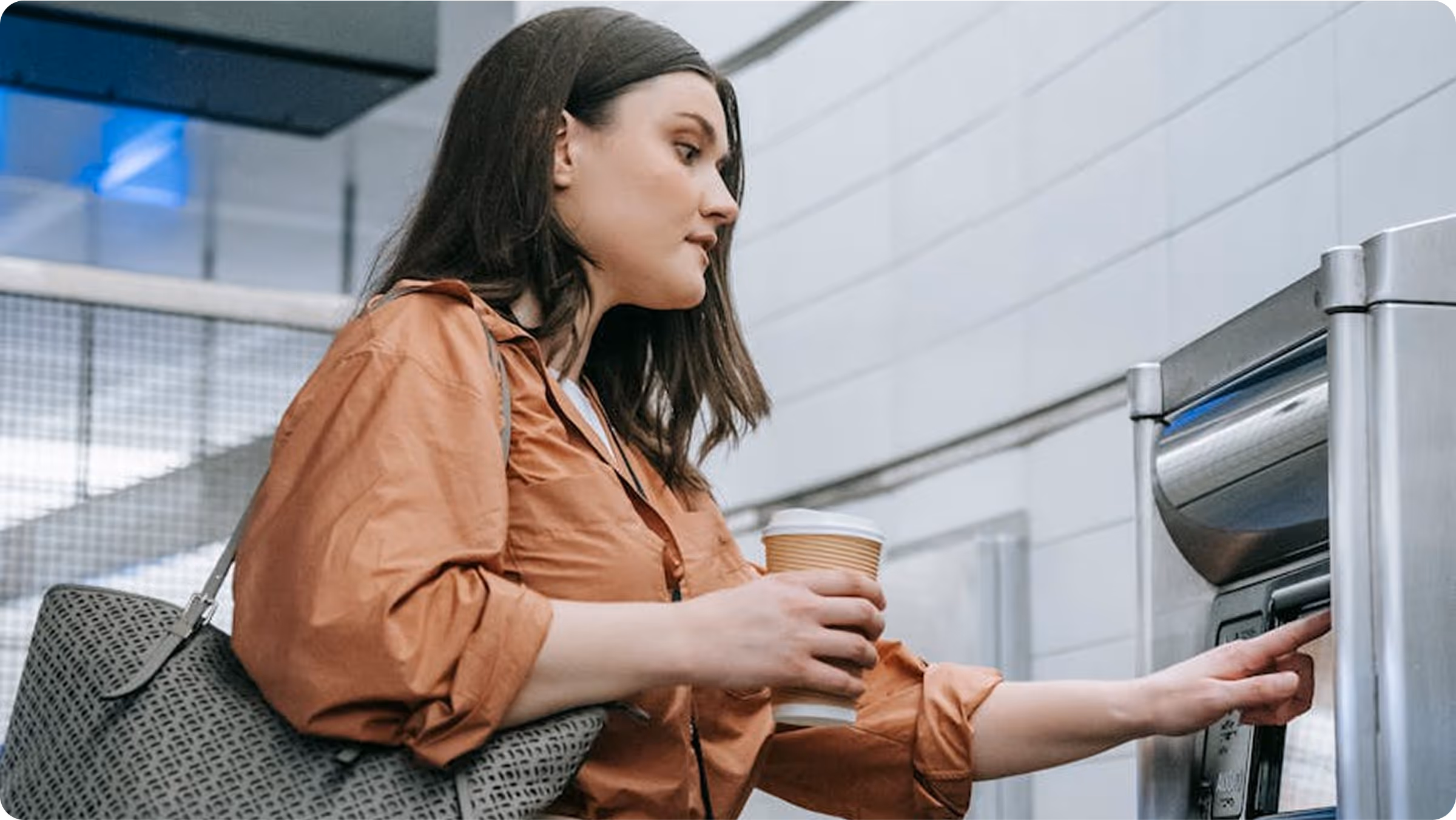 Woman in an orange blouse holding a coffee cup and pressing a button on a metallic parking ticket machine.